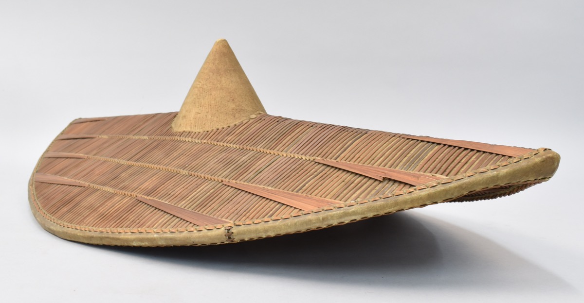 Shield made of wood and cane resting on table. Shield is wooden pointed-oval structure with central ridge, v-shaped cross-section and central conical boss; covered with woven cane strips and strip of hide affixed around rim.